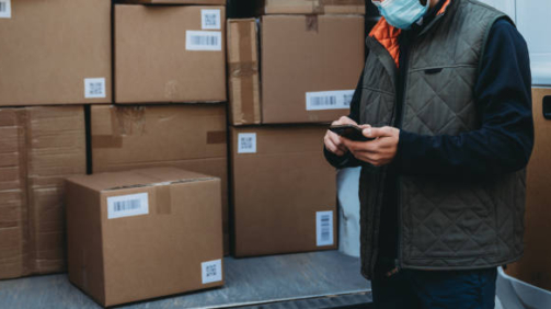 Close up view of a delivery man while he's checking his smart phone in front of the delivery van. He's wearing a protective face mask. Dummy QR code and barcode.